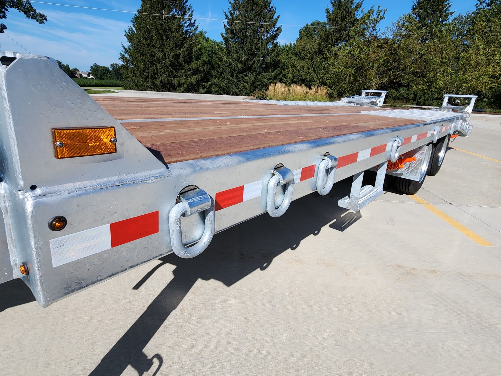 Close view from the left side of a Sauber Mfg. Co. 1580-BTC Beavertail Trailer highlighting the Swivel D-Ring set mounted to the trailer deck perimeter frame. The trailer is shown on concrete in front of an evergreen tree line and blues sky on a sunny day.