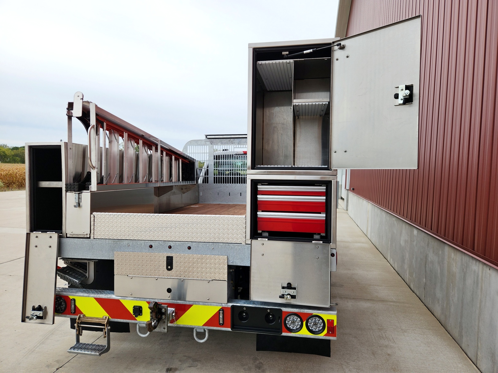 View from rear of a Sauber Mfg. Co. Sign Truck Galvanized Platform Crane Body with Galvanized Bulkhead, Angelim Flooring, Stainless Steel Toolboxes and Step Ladder Storage. Boxes. Custom designs provide storage for sign maintenance tools and materials that you need. The truck is shown in front of a red metal building on concrete.