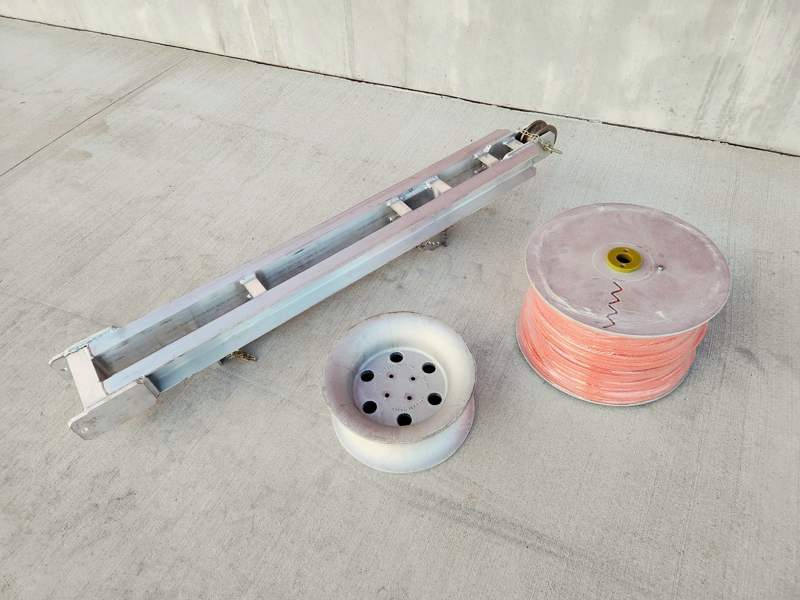 Model 1570-B Puller Underground accessories shown from above on a concrete pad with a concrete foundation in the background. The accessories are a jib arm, 5000lbs capacity capstan and a aluminum cassette with orange underground rope.