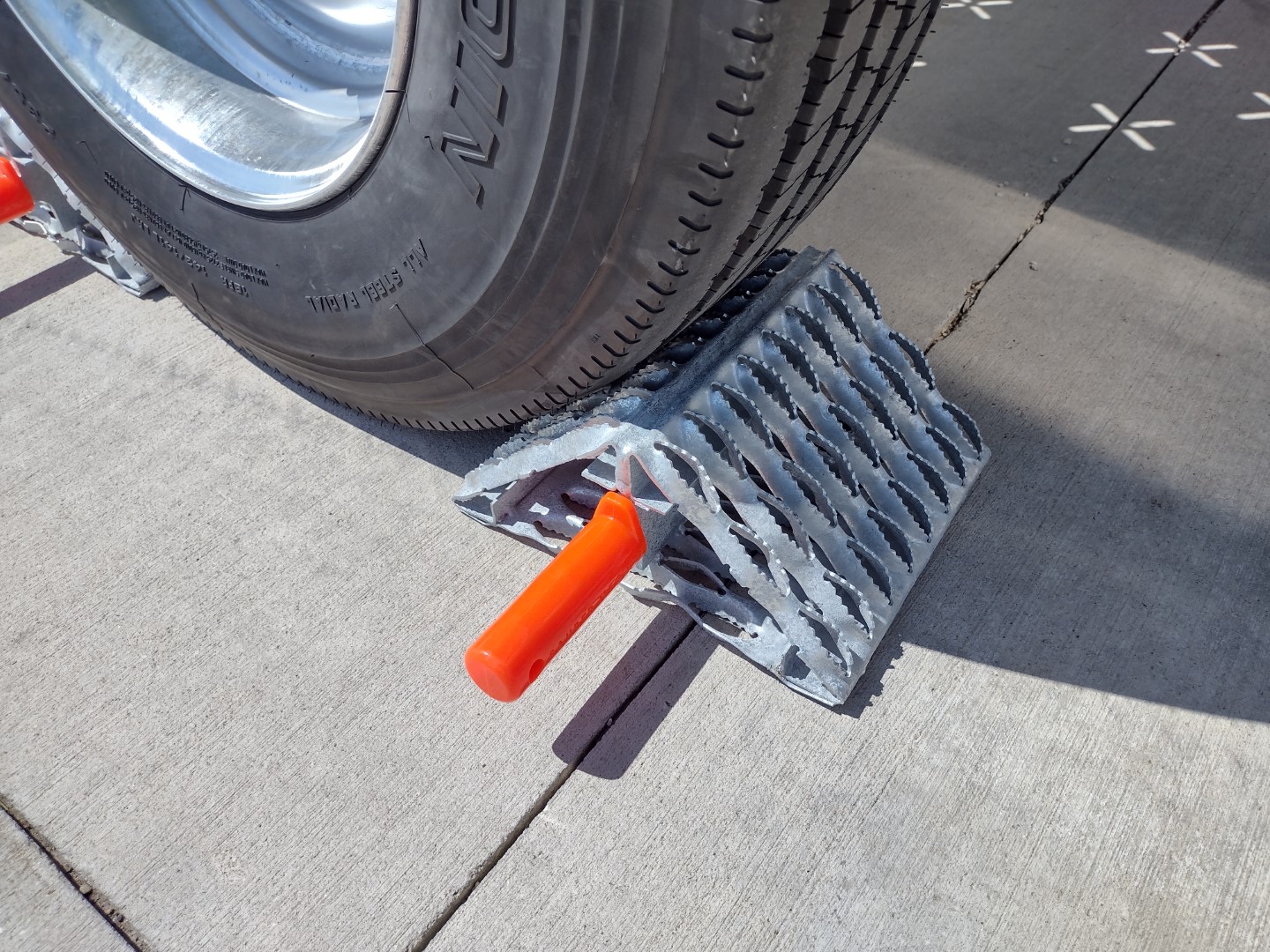 One All-Weather Wheel Chock with steel grip strut surfaces in position on the ground against a tire. Only the wheel and the wheel chock are visible. The All-Weather Wheel Chock has a natural galvanized finish and includes an orange urethane grip handle.