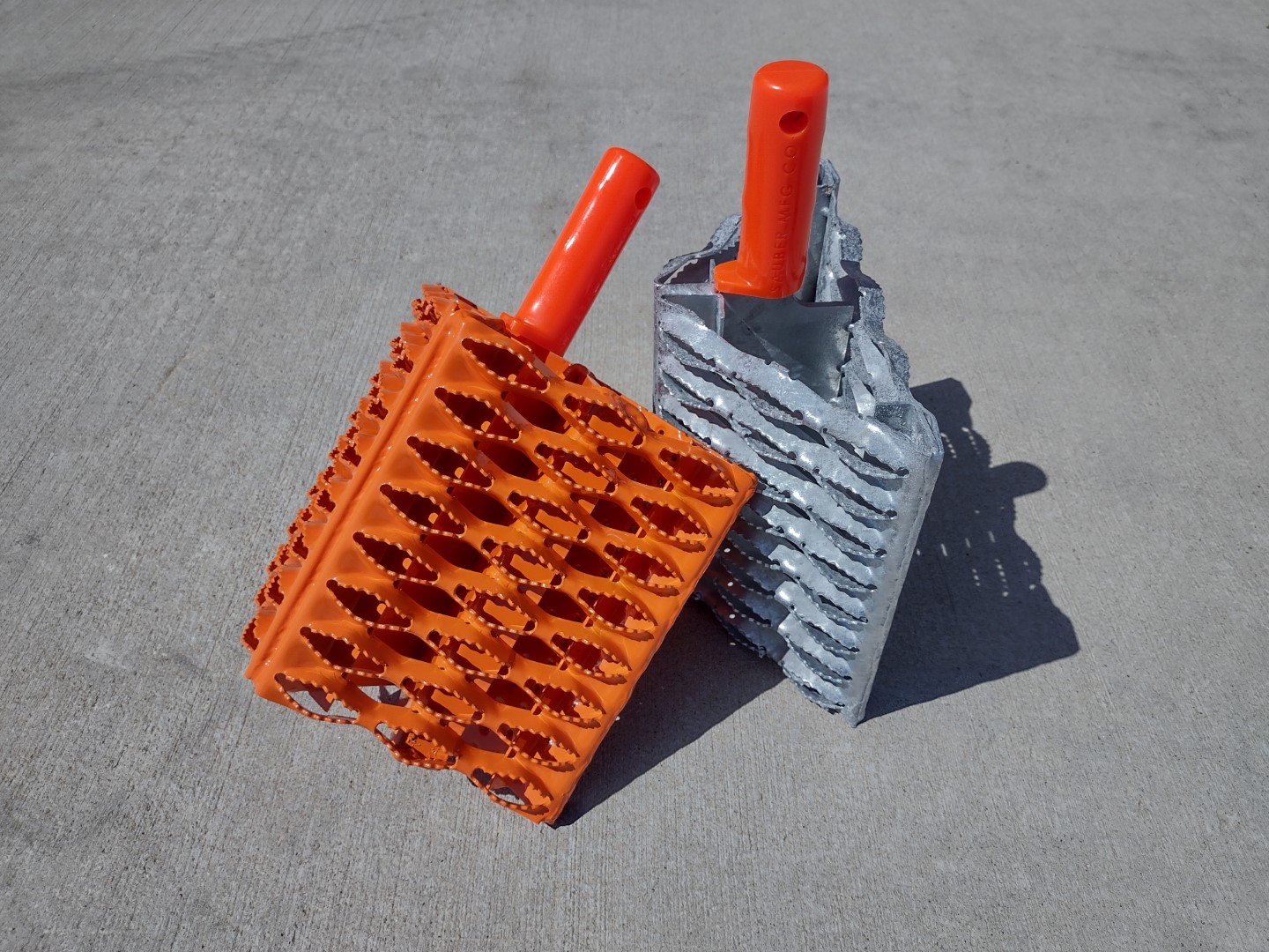 Two Galvanized Steel Wheel Chock with grip strut surfaces leaning against one another. One chock has natural galvanized finish. The other chock is powder coated florescent orange. These chocks have an orange urethane grip handle.