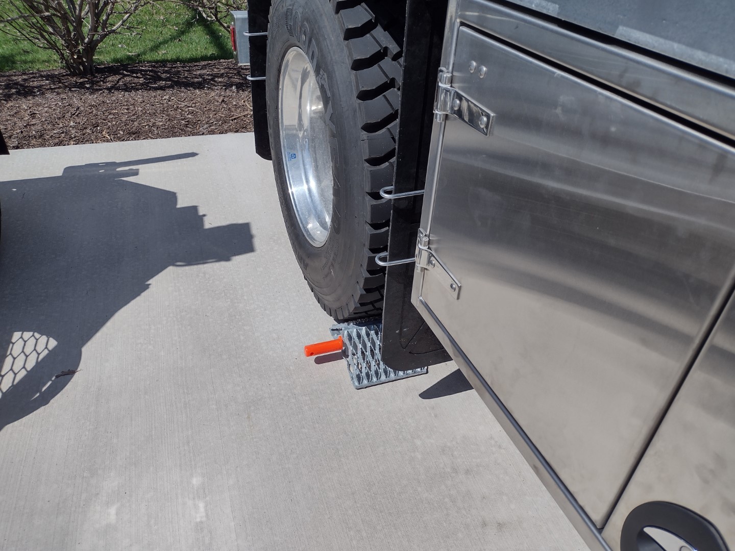 One All-Weather Wheel Chock with steel grip strut surfaces in position on the ground against the drive tire of a truck. Only part of the service body, the wheel and the wheel chock are visible. The All-Weather Wheel Chock has a natural galvanized finish and includes an orange urethane grip handle.
