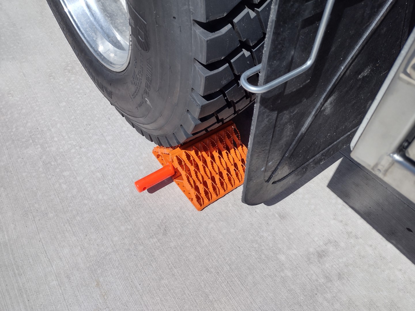 One All-Weather Wheel Chock with steel grip strut surfaces in position on the ground against a tire. The wheel, mudflap and the wheel chock are visible. The All-Weather Wheel Chock has powder coat orange over galvanized finish and includes an orange urethane grip handle.