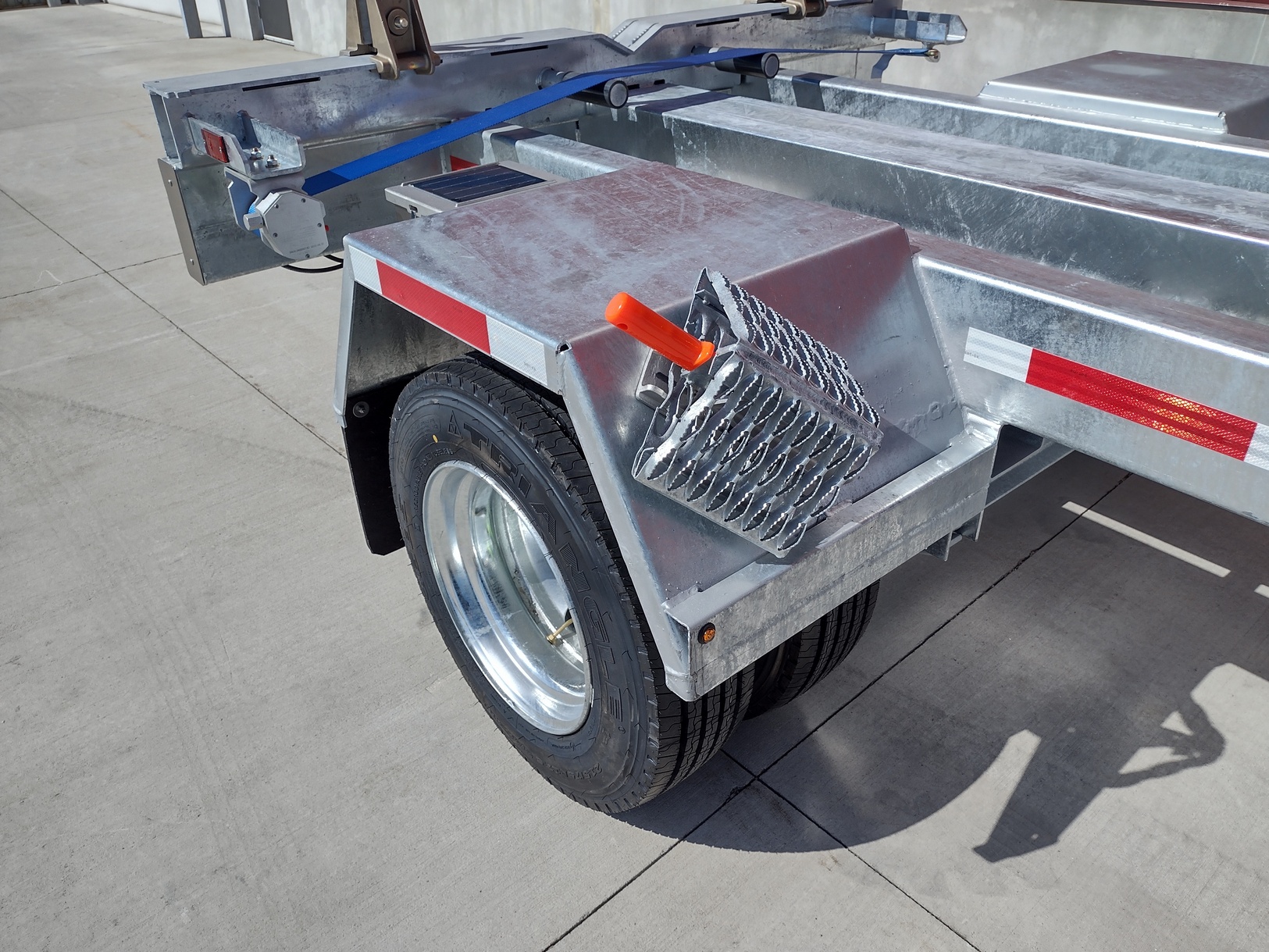 Almag wheel chock holder with Sauber Mfg. Co. All Weather Wheel Chock mounted to trailer fender. The trailer is a hot dipped galvanized Sauber Mfg. Co. Pole Trailer with dual wheels. The view is from the front of the wheels toward to the rear of the trailer. The the fender, one set of dual whees and the trailer rear are visible. Wheels are galvanized finished.
