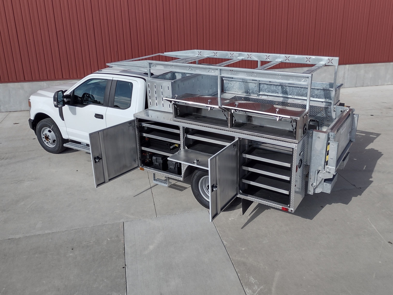 Left side view slightly from the rear of a Sauber Mfg. Co. NexGen Aluminum Service Body. Shown in natural aluminum with stainless steel rear tailgate, stop/turn/backup taillights and lighted recessed ABS plastic license plate holder. Left side compartment top ladder rack is visible. Left side compartments are open showing shelves and storage areas. The truck is shown in front of a red metal building on concrete.