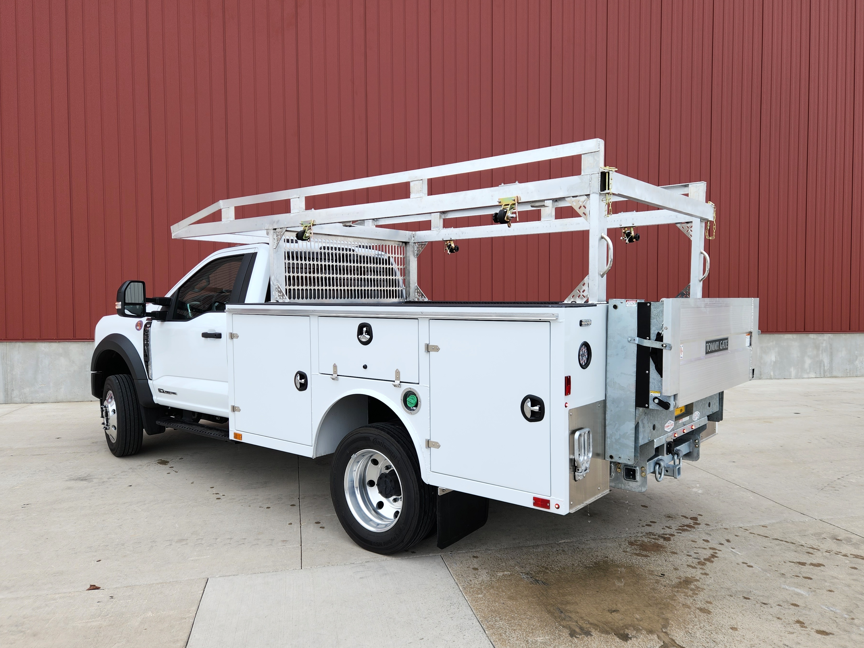 Left side rear view of a Sauber Mfg. Co. NexGen Aluminum Service Body. Shown in white with natural aluminum ladder rack and galvanized lift gate / bumper assembly with hitch. The truck is shown in front of a red metal building on concrete