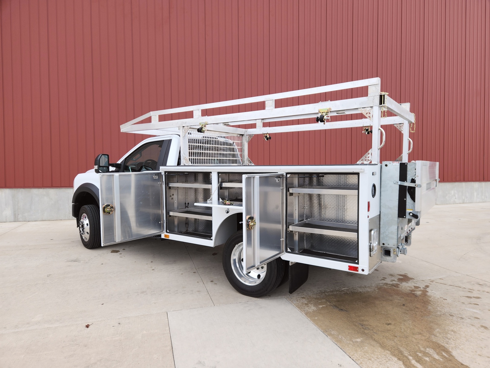 Left side rear view of a Sauber Mfg. Co. NexGen Aluminum Service Body. Shown in white with natural aluminum ladder rack / cab guard and galvanized lift gate / bumper assembly with hitch. Compartment doors are open showing shelves, parts bins and storage. The truck is shown in front of a red metal building on concrete.