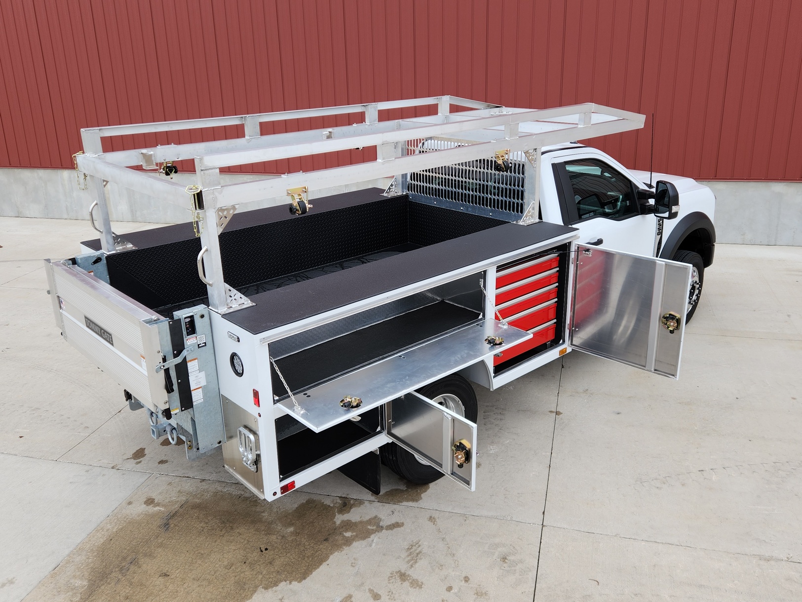 Right side rear view of a Sauber Mfg. Co. NexGen Aluminum Service Body. Shown in white with natural aluminum ladder rack and galvanized lift gate / bumper assembly with hitch. Compartment doors are open showing shelves, parts bins and storage. The truck is shown in front of a red metal building on concrete.
