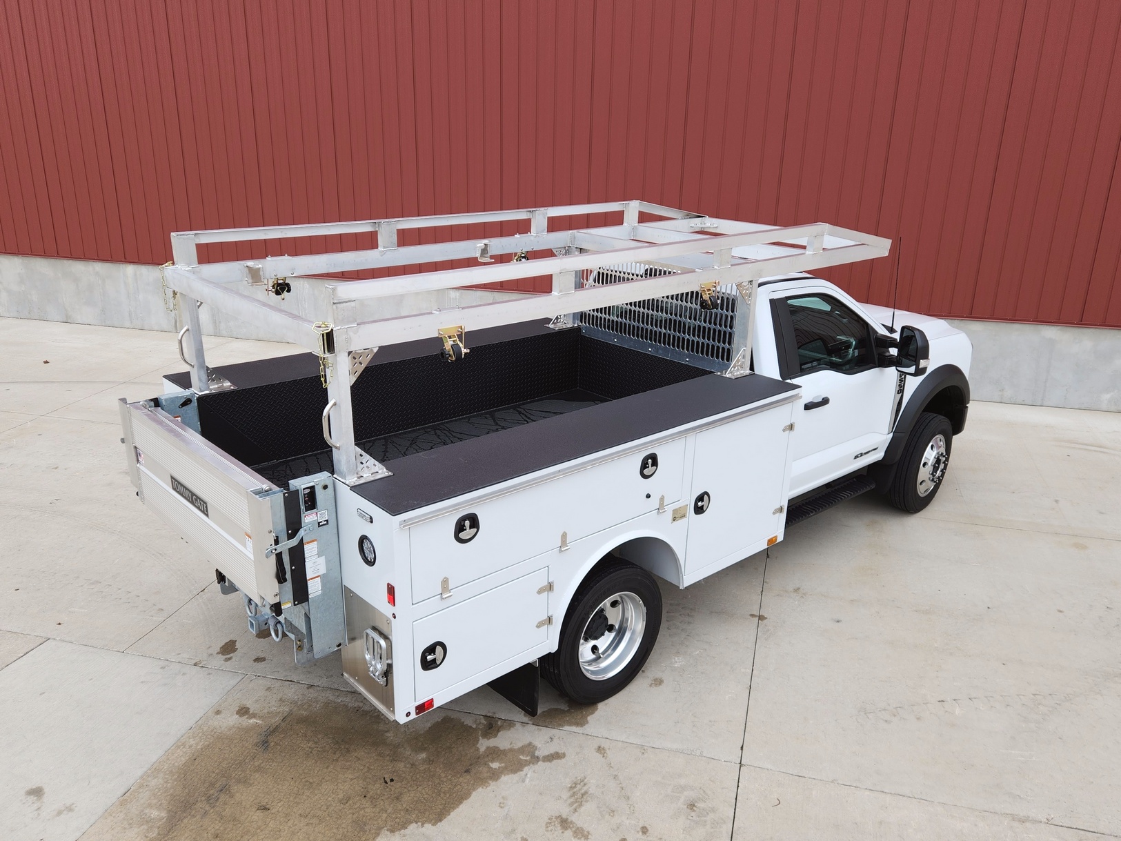 Right side rear view of a Sauber Mfg. Co. NexGen Aluminum Service Body. Shown in white with natural aluminum ladder rack and galvanized lift gate / bumper assembly with hitch. The truck is shown in front of a red metal building on concrete