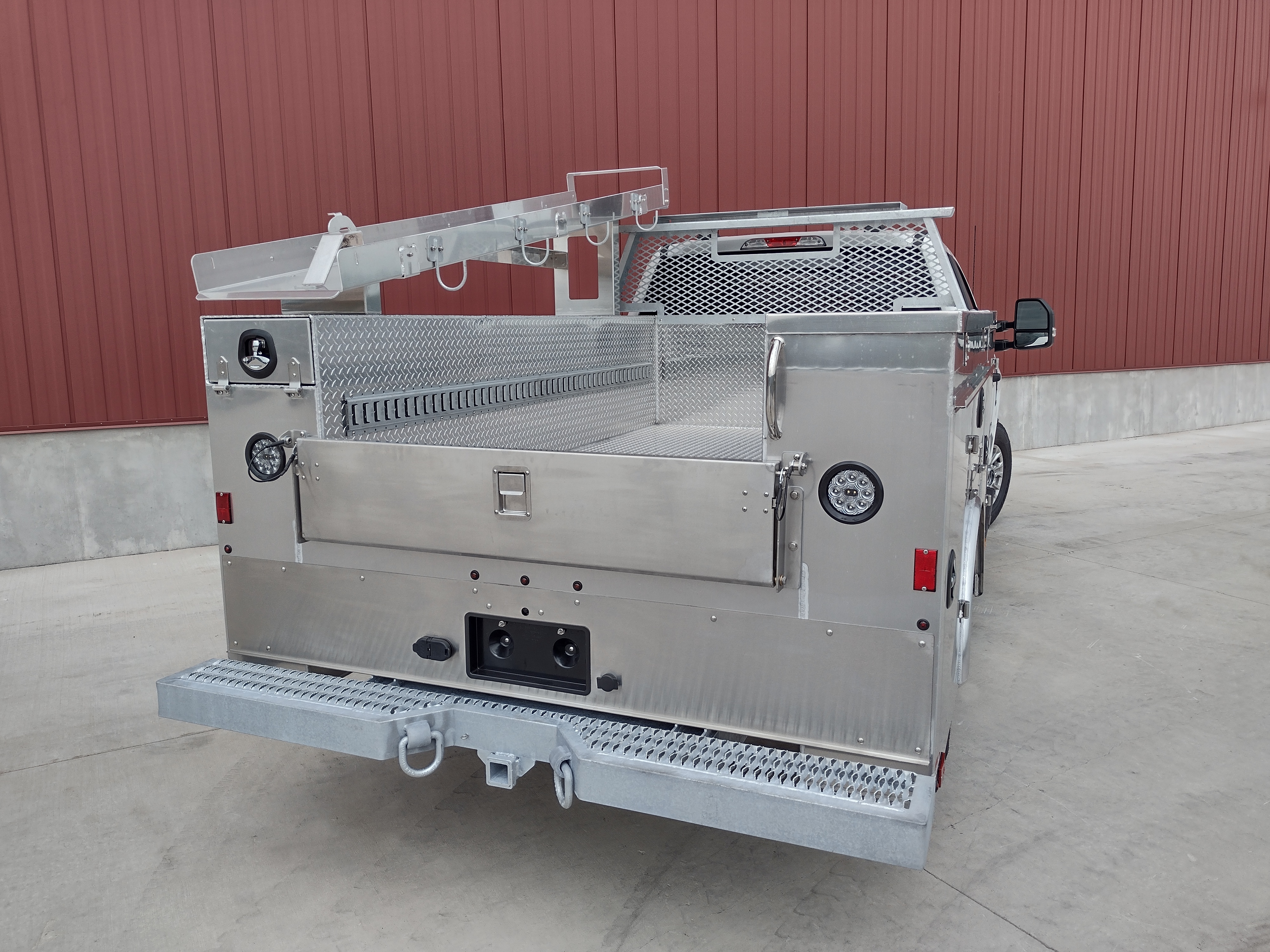 Rear view slightly from the right side of a Sauber Mfg. Co. NexGen Aluminum Service Body. Shown in natural aluminum with stainless steel rear tailgate, stop/turn/backup taillights and lighted recessed ABS plastic license plate holder. Galvanized step bumper with towing provisions. The truck is shown in front of a red metal building on concrete.