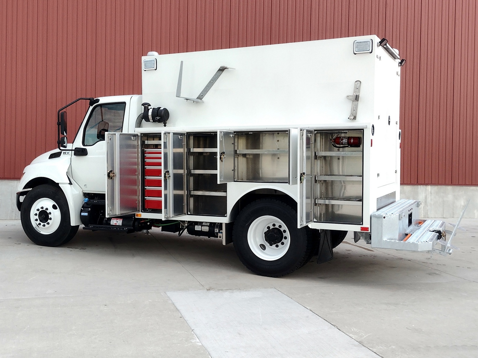 Left side and slightly from the rear view of a Sauber Mfg. Co. NexGen Aluminum Service Body. White with superstructure enclosure on a larger truck with dual axles. Four service compartment doors are open with shelves and tool storage are visible. Galvanized step bumper with safety provisions. The truck is outside and shown in front of a red metal building wall on concrete.