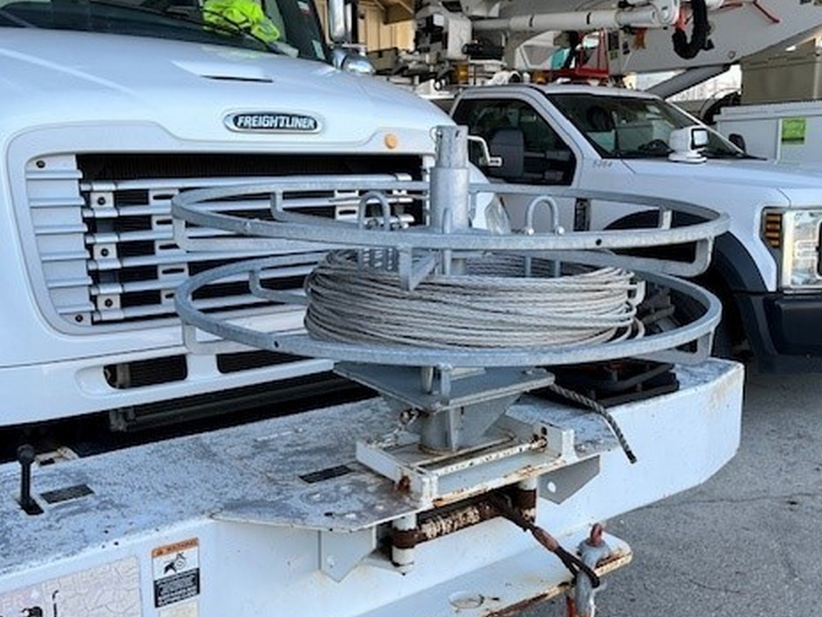 Front right image of a front bumper mounted Sauber Mfg. Co. Hand Coil Reel with adjustable inner diameter and hot dipped galvanized finish. The mounting bracket allows the reel to rotate 90 degrees for easier loading. The part is shown on a line truck bumper with other equipment in the background.