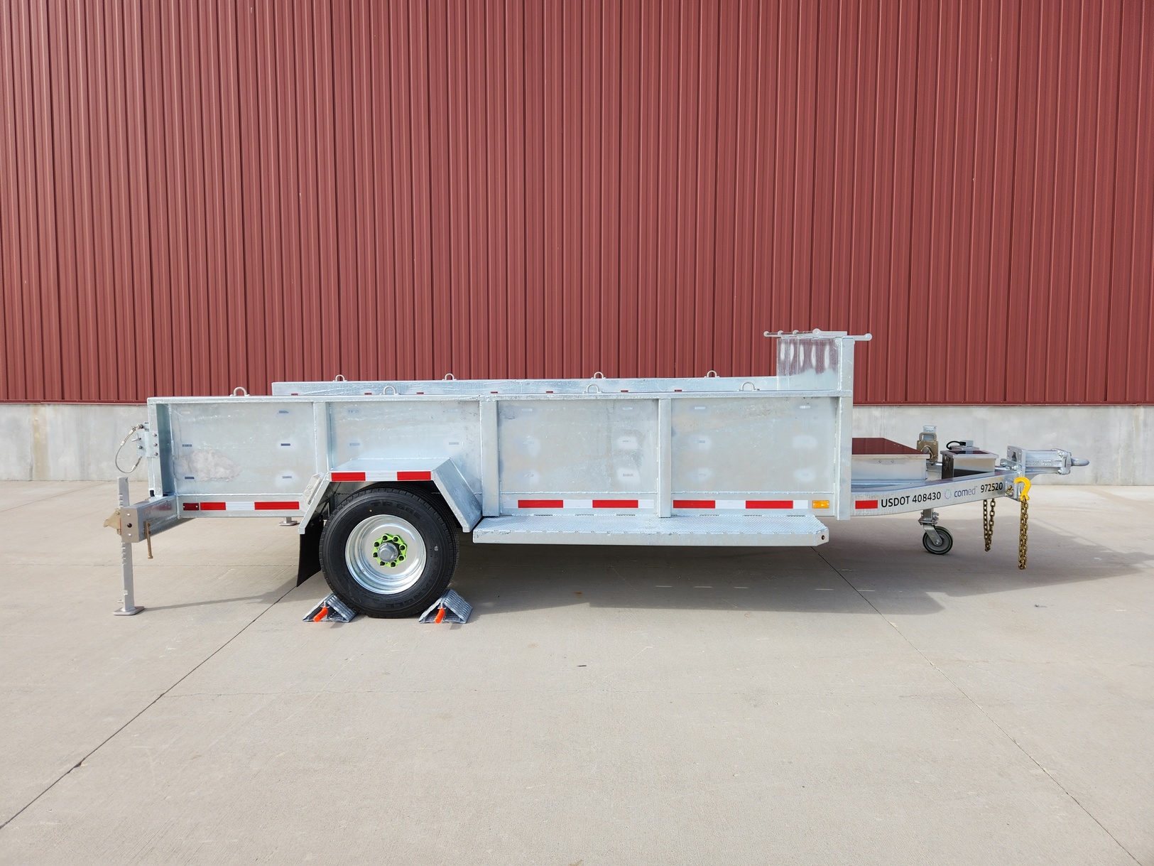 Right side view of a Sauber Model 1560 Reel Reel Cargo Trailer with galvanized finish, bulkhead material rail and stainless-steel a-frame toolbox. It is shown on concrete in front of a red metal building.
